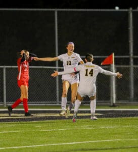 Soccer players celebrating a goal on the field at night, with one opposing player in the background.