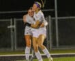 Two soccer players in white uniforms celebrate with a joyful hug on a field at night.