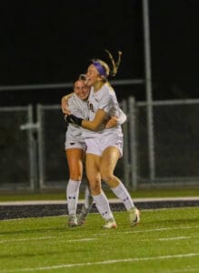 Two soccer players in white uniforms celebrate with a joyful hug on a field at night.