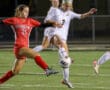 Girls' soccer match at night with player in white kicking the ball, opposed by a player in red.