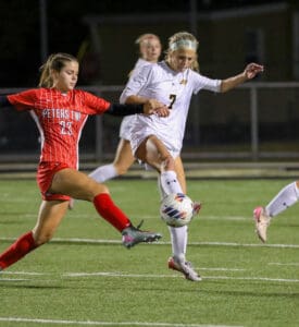 Girls' soccer match at night with player in white kicking the ball, opposed by a player in red.