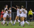 Soccer team in white jerseys celebrates a victory on the field at night, joyful expressions and group hug.