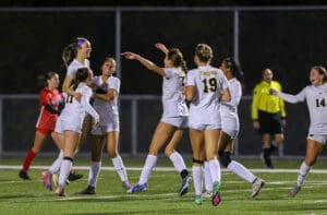 Soccer team in white jerseys celebrates a victory on the field at night, joyful expressions and group hug.