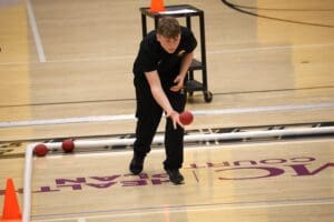Person playing boccia on an indoor court, focused on throwing a red ball towards a marked area.