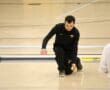 Person in black uniform playing indoor bocce, kneeling on wooden court, ready to roll a yellow ball towards green ones.