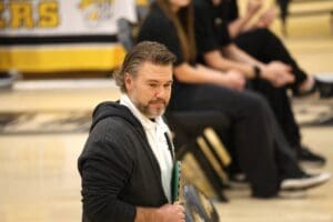 Man in a casual sports outfit holding clipboard at indoor sports event, blurred background of people seated on chairs.