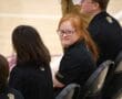 Smiling girl in black sports uniform seated at an indoor event, surrounded by others.