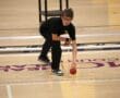 Person bending to pick up a red ball on an indoor court, wearing a black outfit and glasses.