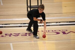 Person bending to pick up a red ball on an indoor court, wearing a black outfit and glasses.