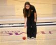 Person playing bocce indoors, rolling a red ball on a gym floor.