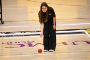 Person playing bocce indoors, rolling a red ball on a gym floor.