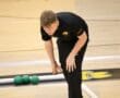 Athlete focused on a bocce game, bending down on an indoor court with green balls nearby.