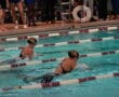 Two competitive swimmers racing in a pool during a swim meet, with spectators watching from the poolside.