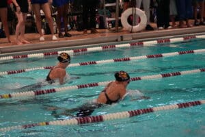 Two competitive swimmers racing in a pool during a swim meet, with spectators watching from the poolside.
