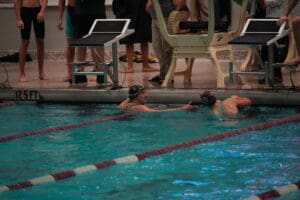 Swimmers at the pool edge during a swim meet, surrounded by starting blocks and spectators.