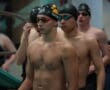 Competitive swimmers wearing swim caps and goggles, preparing for a race at an indoor pool event.