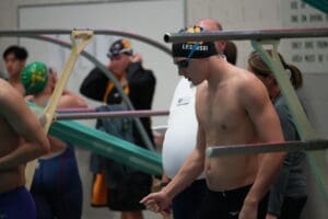 Swimmer prepares before a race, wearing a swim cap and goggles, surrounded by other athletes and coaches.