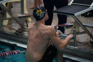 Swimmer prepares for race start at poolside, wearing black swim cap and goggles, gripping starting block.