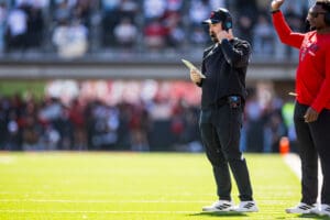 Football coach on the sidelines with headset, holding a play card, during a sunny game day.