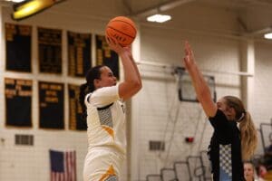Basketball player in white jersey jumps to shoot, defended by player in black jersey in a gymnasium.
