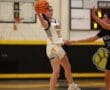 Girls' basketball game action, player in midair pass against defender on school gym court.