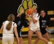 Girls' basketball game action with player in white holding ball, facing defenders in black jerseys on the court.