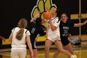 Girls' basketball game action with player in white holding ball, facing defenders in black jerseys on the court.
