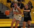 Basketball game in action: Player in white dribbles past opponent in black at indoor court with yellow seating.