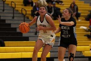 Basketball game in action: Player in white dribbles past opponent in black at indoor court with yellow seating.