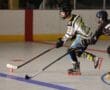 Two roller hockey players compete intensely on the rink, one in black and the other in white and yellow, focused on the puck.