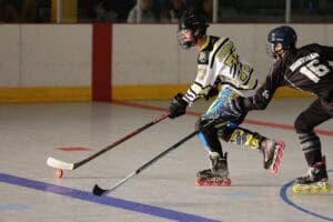 Two roller hockey players compete intensely on the rink, one in black and the other in white and yellow, focused on the puck.