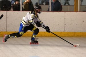 Roller hockey player in action, wearing a white jersey, maneuvering the puck on the rink with spectators in the background.