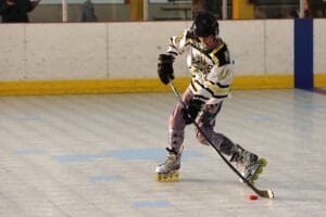 Roller hockey player in action, skating with puck on indoor rink, wearing black helmet and yellow jersey.