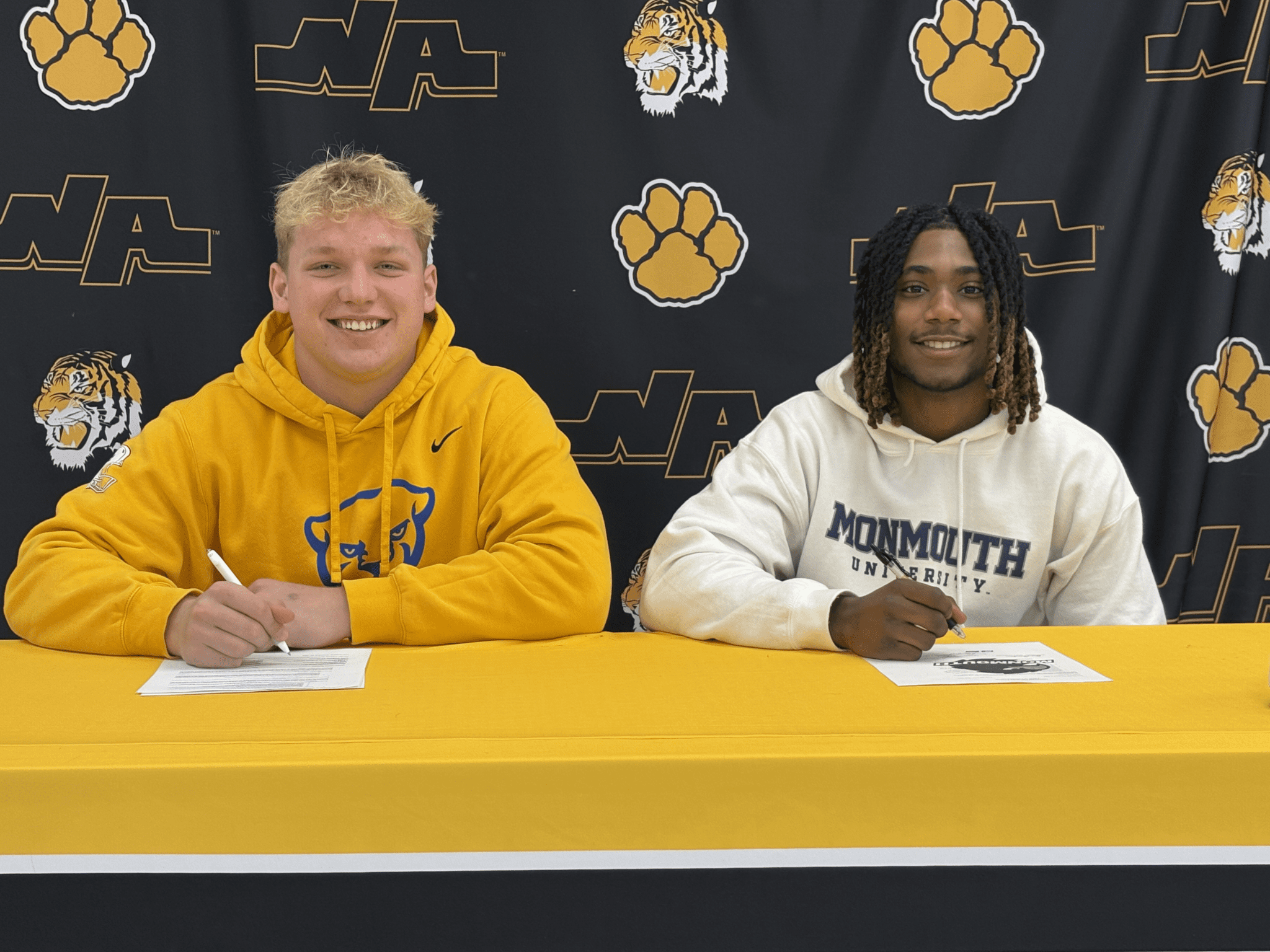 Two students smiling while signing documents at a table, wearing university sweatshirts, with a tiger backdrop.