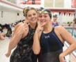 Two female swimmers smiling and posing in swimsuits by an indoor pool.