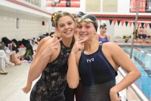 Two female swimmers smiling and posing in swimsuits by an indoor pool.