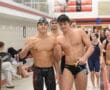Swimmers posing at an indoor pool event, wearing swim caps and gear, with teammates in the background.