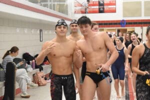 Swimmers posing at an indoor pool event, wearing swim caps and gear, with teammates in the background.