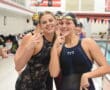 Two smiling female swimmers pose with peace signs by the pool in swim gear.