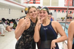 Two smiling female swimmers pose with peace signs by the pool in swim gear.
