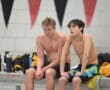 Two male swimmers rest poolside, surrounded by towels and water bottles, in a tiled indoor swimming facility.
