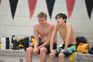 Two male swimmers rest poolside, surrounded by towels and water bottles, in a tiled indoor swimming facility.