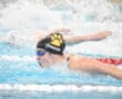 Swimmer in goggles and cap competes in butterfly stroke at indoor pool, creating splashes.