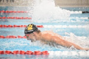 Swimmer performing butterfly stroke in pool, wearing black swim cap with yellow paw logo.