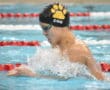 Swimmer in action at indoor pool, wearing swim cap and goggles, splashing water during competition.