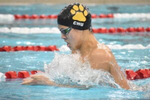 Swimmer in action at indoor pool, wearing swim cap and goggles, splashing water during competition.
