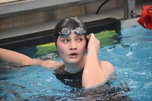 Teen swimmer adjusting goggles at the pool edge in black swim cap and suit.