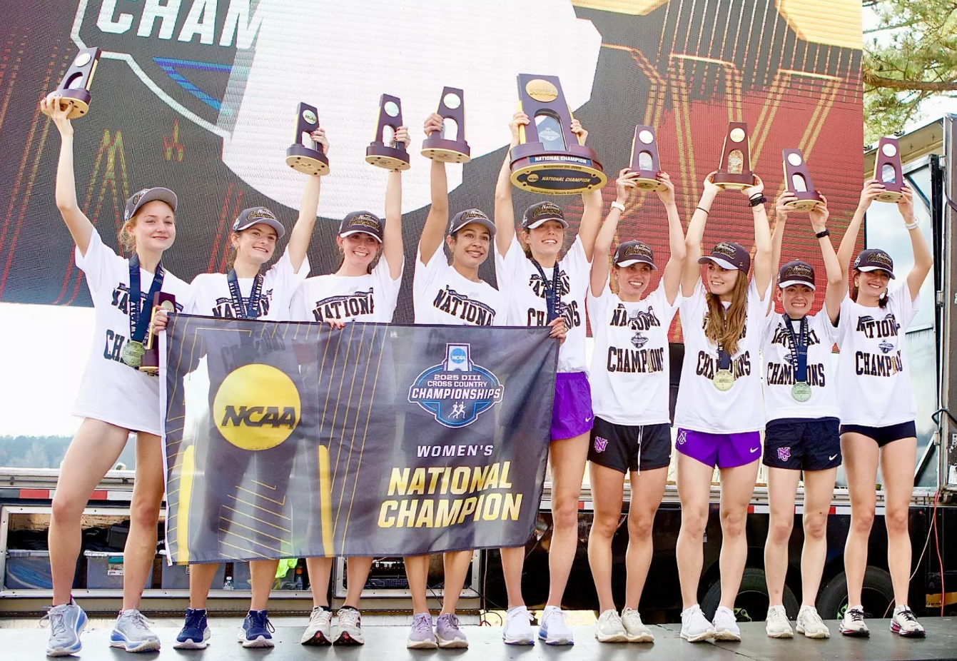 Women's cross country team celebrates NCAA Championship victory with trophies and banner.
