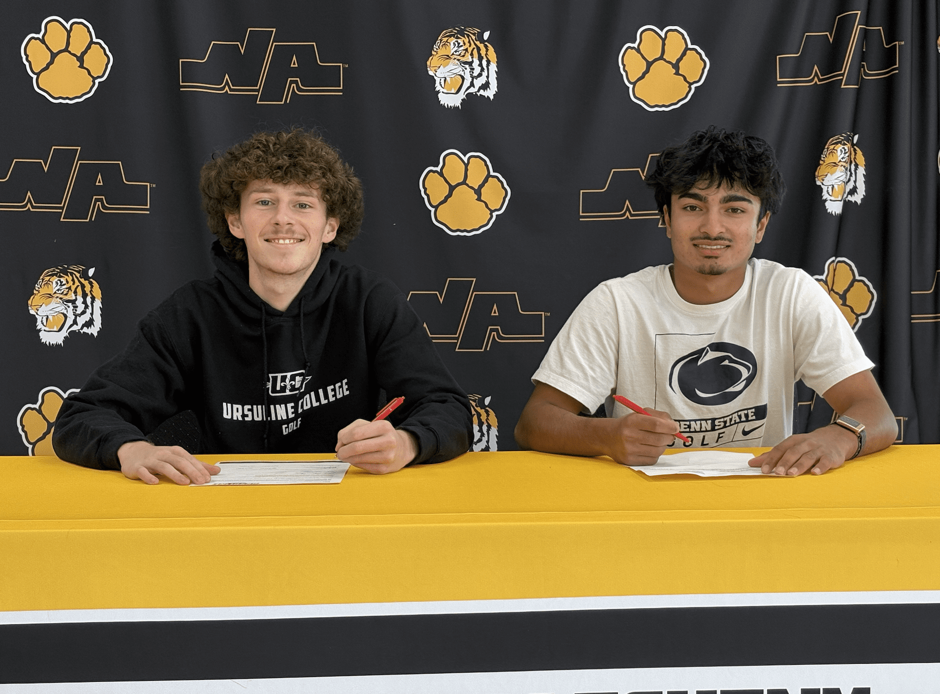 Two young men signing documents at a table, with school logos in the background.