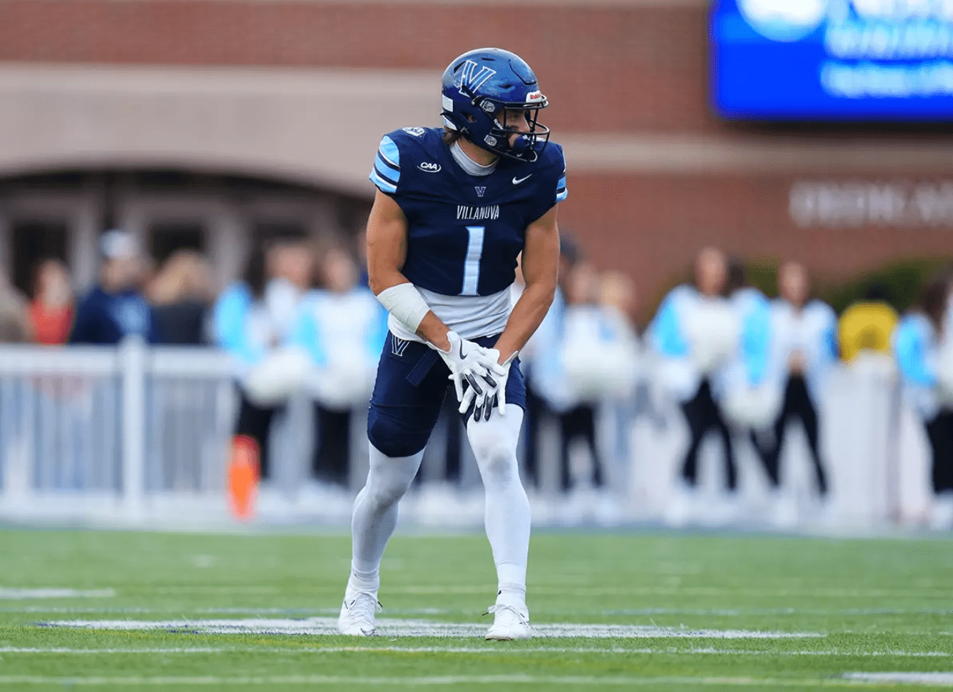 Football player in Villanova uniform ready on the field during a game, with focused cheerleaders in the background.